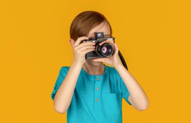 Kameralı çocuk. Genç çocuk klasik bir kameradan fotoğrafçılık mesleğini deniyor. Sarı arka planda stüdyo portresi. Kamera kullanan bir çocuk..
