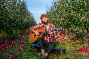 Kareli tişörtlü sakallı bir adam gitar çalar ve yeşil çimenli bir parkın arka planına gülümser. Açık hava eğlencesi, hafta sonu. Elma bahçesinde dinlenen gitarlı yakışıklı genç adam..