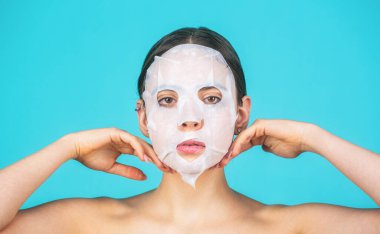 Yüzünde nemlendirici maske olan bir kadın. Kadın yüzüne pamuk yüz nemlendirici maske takıyor, cildine iyi bakıyor, kırışıklıkları önlüyor, izole edilmiş mavi arka planda duruyor..