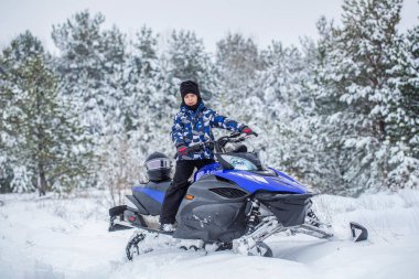 Kış manzarasında kar arabası süren bir çocuk. Karlı bir havada, ormanda, kar arabasında gülümseyen küçük bir çocuk. Karlı bir patikada, çam ağaçlarında, mavi bir kar aracına binen çocuk..