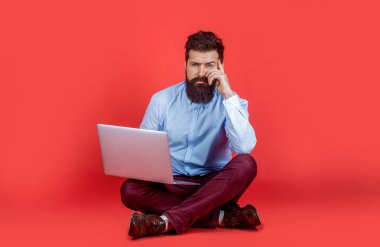Dizüstü bilgisayarda çalışan adam. Sakallı adam kırmızı arka planda dizüstü bilgisayarla yerde oturuyor. Dizüstü bilgisayarı tutuyorum. Bilgisayarlarını kullanan bir iş adamı. Ciddi sakallı işçi dizüstü bilgisayar.