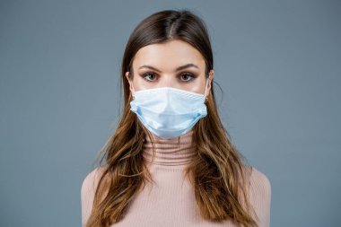 Tıbbi maske takan bir kadın. Corona virüsü için ameliyat maskesi takan bir kadın. Yüzü maskeli kız, gri duvarın üzerinden dışarı bakıyor..