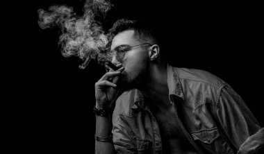 Gözlüklü ve siyah arka planda puro içen bir adam. Etrafında dönen duman, yanan ateşi yakıyor. Siyah ve beyaz.