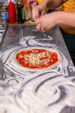 Şef pizzaya peynir serper. Yakın plan, eller pizza hamuruna domates soslu peynir serpecek. Mutfakta pizza yapan şef fırıncının eli. Adam masada pizzaya peynir ekliyor..