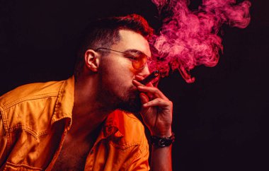 Etrafında dönen duman, yanan ateşi yakıyor. Gözlüklü ve puro içen adam, sigara ve dumana karşı..