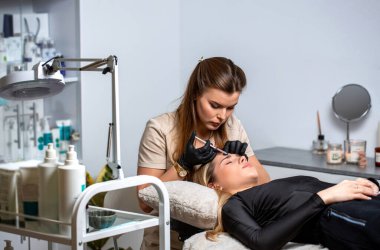 Beyaz önlüklü ve koruyucu eldivenli doktor kadın hastaya alnına tıbbi iğne yapıyor. Hastane kliniğinde osmetik cerrahi bölümü. Alnına enjeksiyon yapılan genç bir kadın..