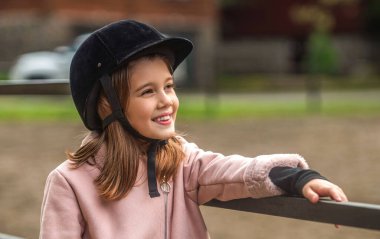 Küçük kız ahırda gülen bir jokey kaskı. Jokey giymiş bir kız çocuğunun portresi bir at biniciliği eğitimi.