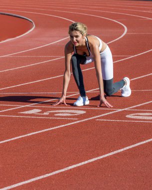 Sporcu kadın başlangıç çizgisinde çömelmiş kırmızı koşu pistinde koşmaya hazır. Spor giyimli koşucu kadın kırmızı kaplamalı stadyum pistinde koşmaya hazırlanıyor..