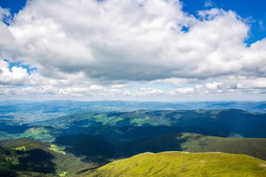 Yüksek tepeleri, yeşil tepeleri ve vadide alçak bulutları olan dağ manzarası. Yeşil çayırlar ve karla kaplı dağlardaki manzara. Dağlar yüksek kayalık zirveler.