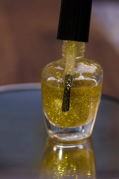 A bottle of translucent gold glitter nail polish on a mirror surface
