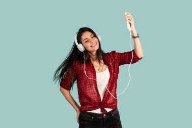 Çizgili tişörtlü neşeli, esmer kadın pastel mavi arka planda yalnız poz veriyor. Kulaklıkla müzik dinlemek ve dans etmek