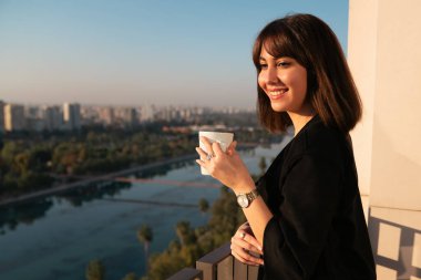 Genç bir kadın gülümsüyor, kahve içiyor ve balkondan dışarı bakıyor. Yüksek kalite fotoğraf
