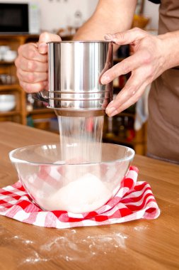 Kişi yavaş yavaş cam bir kase içine süzgeçten un ekliyor. Mutfakta lezzetli bir tarifin bileşenini hazırlıyor.