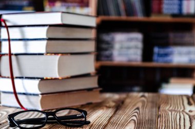 Siyah gözlükler ve eski kitaplar arka planda bir kitaplıkla ahşap bir masanın üzerinde duruyor.