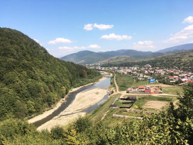 Yazın dağlarda doğal manzara. Parlak mavi gökyüzü ile güneşli kırsal manzara. Doğa koruma kavramı. Nehir ve küçük evlerle dağ köyünün nefes kesici manzarası.