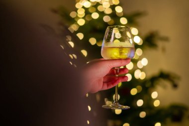 Elinde bir kadeh şarap, arka plandaki yılbaşı ağacında bulanık noel ışıkları. Parlak çam ağacıyla süslenmiş, elinde bardakla içki tutan biri..