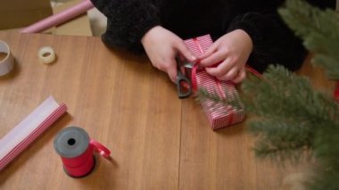 Kırmızı kurdeleyle hediye paketi yapan bir kadın. Eller kırmızı ve beyaz yılbaşı kağıdında bir kutu paketliyor.. 