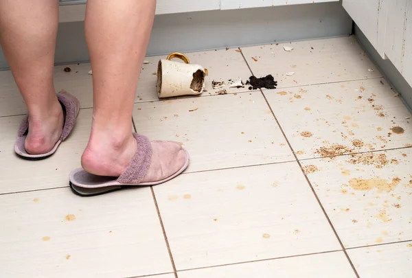 Woman standing next to broken tea cup laying on the kitchen floor, smashed coffee mug and coffee grounds all over the tiles.