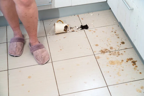 Woman standing next to broken tea cup laying on the kitchen floor, smashed coffee mug and coffee grounds all over the tiles.
