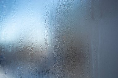 Sisli bir pencere, küçük su damlalarıyla kaplı cam bir yüzey. Camda bulanık bir arkaplan görebilirsiniz, mavi ve gri tonlarıyla. Görüntü serinlik ve nem dolu bir atmosfer taşır.