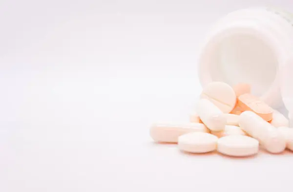 Beyaz bir arka planda, açık beyaz bir kutudan dökülen haplar ve kapsüller gösteriliyor. Farklı şekil ve renklerde ilaçlar: yuvarlak, oval ve kapsüller, çoğunlukla beyaz ve turuncu tonlarda.