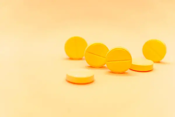 Close-up photo of several round yellow pills on a soft orange background. The pills are scattered in a casual arrangement, with some lying flat and others standing on their edges.
