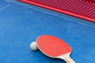 yatay kompozisyonda mavi bir pingpong masasında pingpong raketi, top ve net açısı