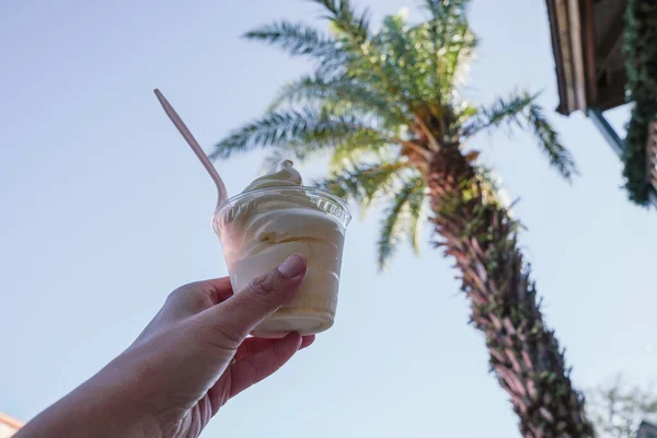 Hand holds up a cup of soft serve pineapple vanilla swirl ice cream, with palm trees in the background