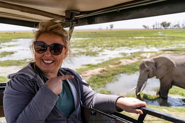 Happy woman on safari watches in excitement as an elephant drinks outside the safari vehicle
