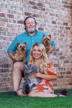 Adult childfree couple with pets - cat and two Yorkies dogs, holding in their arms, against brick wall. Blonde woman, smiling pet friendly clipart