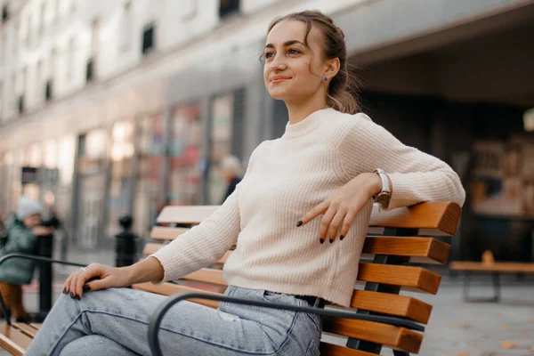 Stock image smiling girl in a knit sweater, sitting on a bench in the city in autumn. Laughing elegant woman chilling after autumn walk.