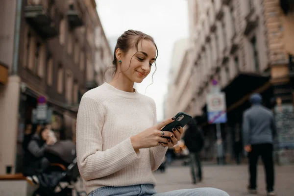 Young stylish woman in sweater using a smart phone standing outdoors on the street in Warsaw.