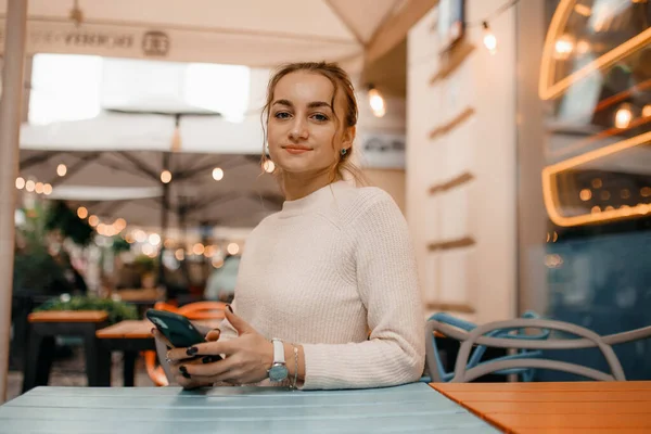 Young stylish woman in sweater using a smart phone standing outdoors on the street in Warsaw.