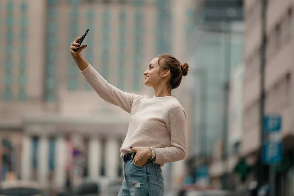 Young woman walking with smart phone on the town square in Warsaw city, Poland. Lifestyle portrait of a young stylish woman in in a beige sweater with smart phone walking on the street in the city.