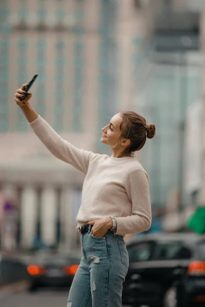 Young woman walking with smart phone on the town square in Warsaw city, Poland. Lifestyle portrait of a young stylish woman in in a beige sweater with smart phone walking on the street in the city.