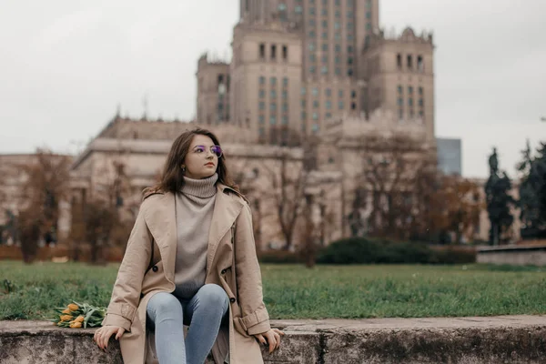 文化宮殿の近くの散歩 ワルシャワの文化宮殿を背景に黄色のチューリップを持つ女の子 花のあるメガネの可愛い女の子がヨーロッパの街を歩く — ストック写真
