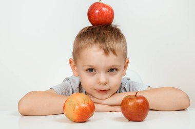 Çocuğun elinde elma, sağlıklı yiyecekler ve vitaminler, gülümseyen, beyaz bir arka plan var. İzole edilmiş beyaz bir arka planda kırmızı elmalı neşeli bir çocuk. Kafasında kırmızı bir elma olan yakışıklı bir çocuğun portresi..