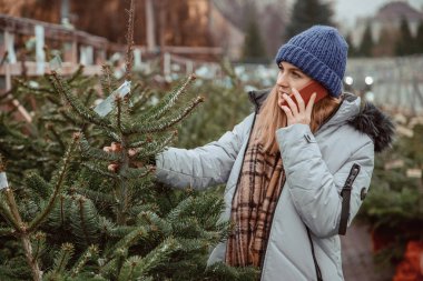 Zarif bir kız Noel ağacı alır. Neşeli kadın Noel ağacının yanında dikilirken telefonda konuşuyor. Genç bir kadın Noel ağacıyla dikilirken telefonu kullanıyor. Yeni yıl ruhu ve perakende fikri.