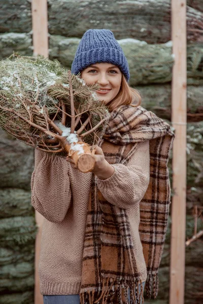 青い帽子の美しい女の子 スカーフは彼女の肩にクリスマスツリーを運びます クリスマスに身を包んだ女の子は クリスマスのために飾られる新鮮な切ったモミの木を家に持ち帰る 緑のコンセプトを守る — ストック写真