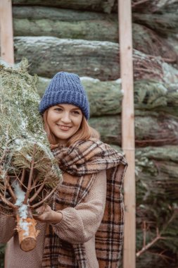 Mavi şapkalı güzel bir kız, atkısı omuzunda bir Noel ağacı taşıyor. Noel 'de giyinmiş bir kız Noel' de süslemek için yeni kesilmiş bir köknar ağacı getiriyor. Yeşil konsepti koru.