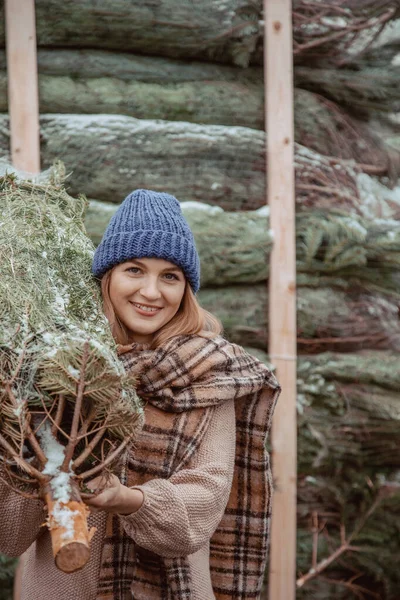 青い帽子の美しい女の子 スカーフは彼女の肩にクリスマスツリーを運びます クリスマスに身を包んだ女の子は クリスマスのために飾られる新鮮な切ったモミの木を家に持ち帰る 緑のコンセプトを守る — ストック写真