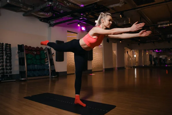 Beautiful young blond girl yoga instructor doing on a mat in a floor standing in the gym. Young sport woman standing on one leg and exercising. Portrait of sport girl doing yoga.