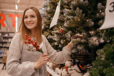 Xmas mağazasında yapay Noel ağacı seçen sevimli genç bir kadın. Noel konsept mağazasında inanılmaz Avrupalı. Bir kadın mağazadan Noel ağacı için yeni yıl süsü alıyor..