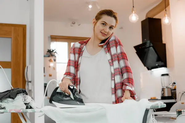 A young woman is ironing clothes and talking on the phone. Sweet pretty housekeeper ironing family clothing at home. Cute Woman ironing clothes in a utility room.