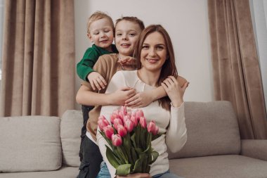 İki gülümseyen çocuğu tarafından kucaklanan pembe lalelerle dolu bir buketi olan bir kadın kanepeye oturur. Sevgi ve aile bağlarıyla dolu iç açıcı bir sahne. Uluslararası Kadınlar Günü için mükemmel. Anneler Günü..