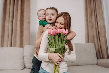 İki gülümseyen çocuğu tarafından kucaklanan pembe lalelerle dolu bir buketi olan bir kadın kanepeye oturur. Sevgi ve aile bağlarıyla dolu iç açıcı bir sahne. Uluslararası Kadınlar Günü için mükemmel. Anneler Günü..