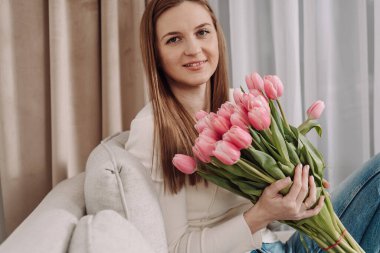 Elinde pembe lalelerle dolu bir buket tutan genç bir kadın, rahat bir koltukta oturuyor. Barışçıl ve düşünceli bir ifade. Yumuşak doğal ışık, modern iç mekan, bahar titreşimleri. 8 Mart. Laleler.