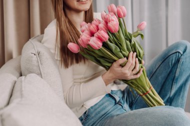 Elinde pembe lalelerle dolu bir buket tutan genç bir kadın, rahat bir koltukta oturuyor. Barışçıl ve düşünceli bir ifade. Yumuşak doğal ışık, modern iç mekan, bahar titreşimleri. 8 Mart. Laleler.