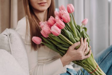 Elinde pembe lalelerle dolu bir buket tutan genç bir kadın, rahat bir koltukta oturuyor. Barışçıl ve düşünceli bir ifade. Yumuşak doğal ışık, modern iç mekan, bahar titreşimleri. 8 Mart. Laleler.