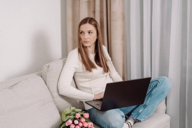 Kanepede dizüstü bilgisayarla oturan ve gözlerini kaçıran gündelik kıyafetli genç bir kadın. Yanında pembe laleler var. Konforlu ev dekorasyonu, ev işi konsepti, modern yaşam tarzı..